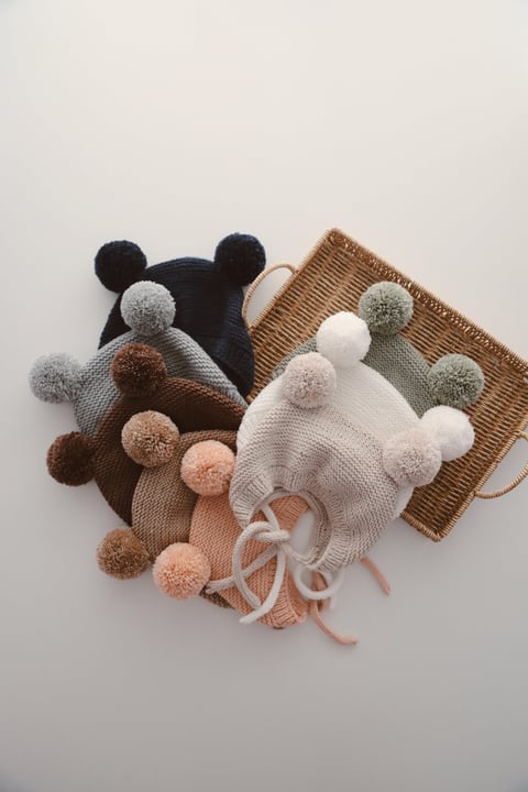 A collection of knitted hats with double pom-poms in a variety of colors, including brown, coffee latte, stone, white, green, beige, salmon, gray, and navy blue